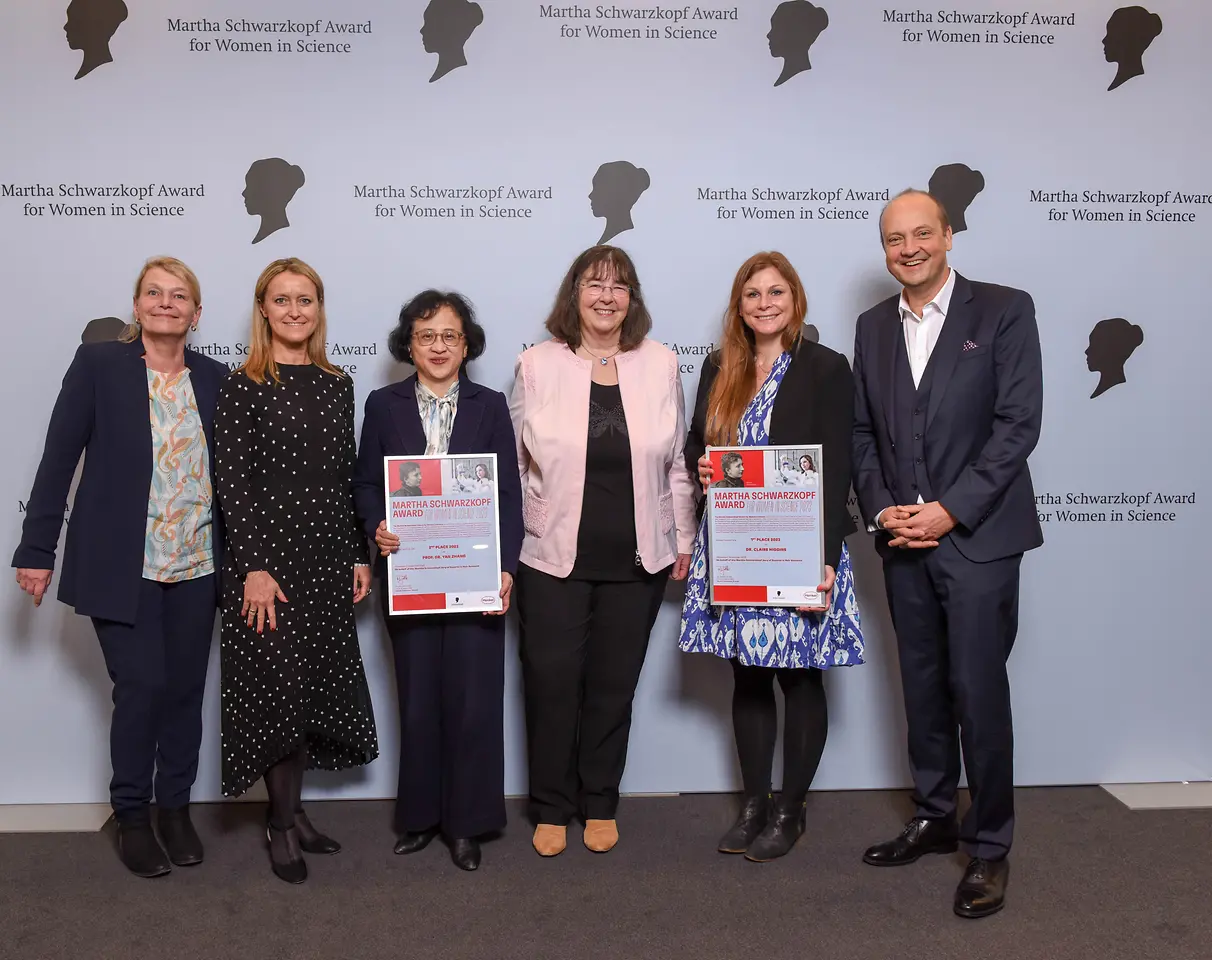 Four Henkel representatives and the two winners of the Martha Schwarzkopf Award are standing next to each other in front of the photo wall. 